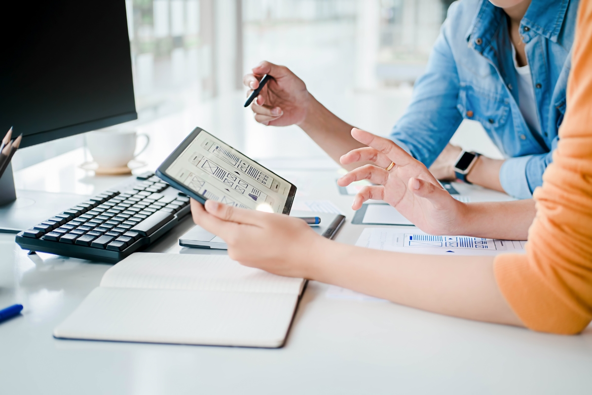 Two people collaborate at a desk with a tablet, keyboard, and notebook. One person holds the tablet displaying a design, while the other gestures towards it with a pen, discussing strategies on how to get featured snippets.