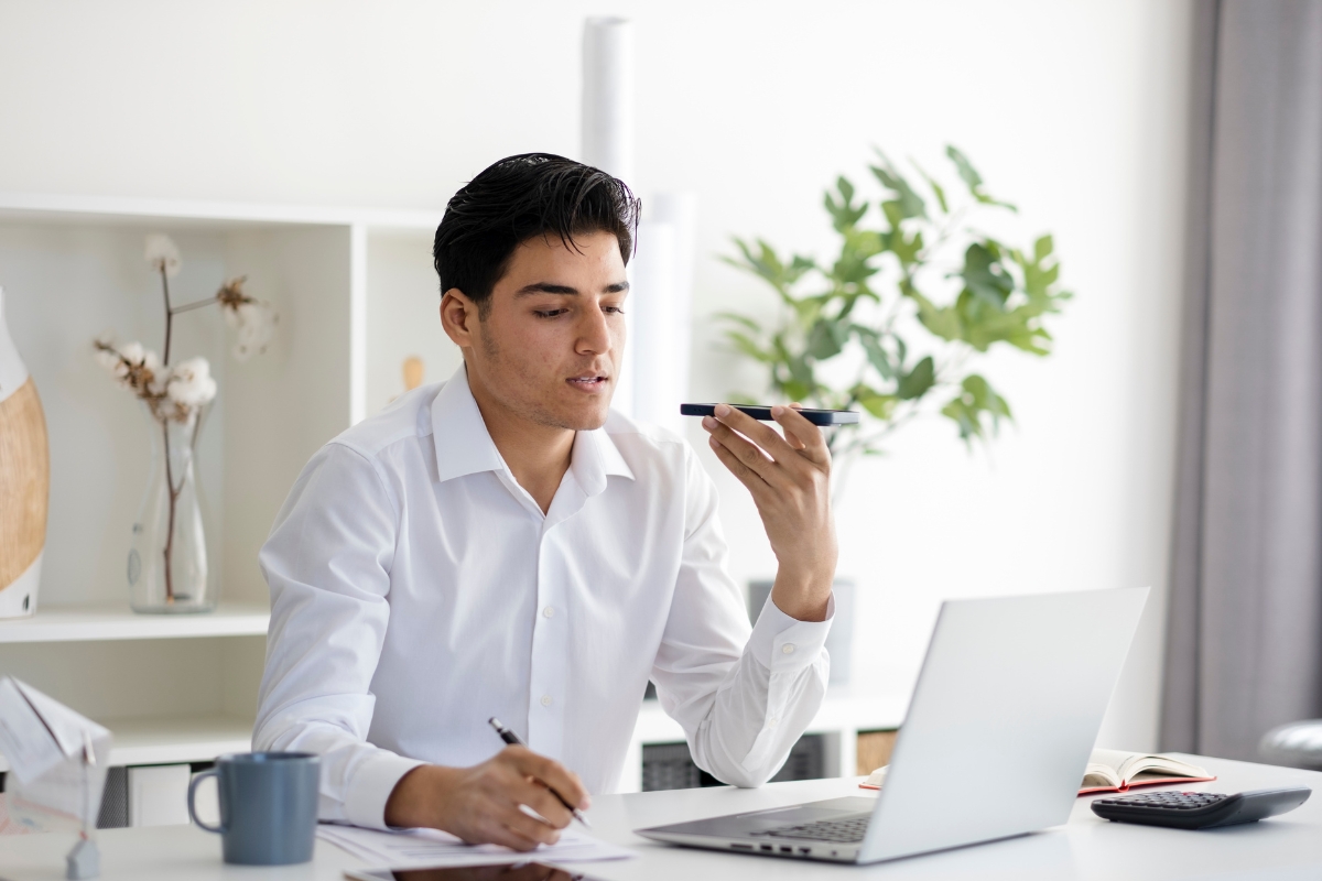 A person in a white shirt is sitting at a desk, speaking into a phone about how to optimize your website for voice search, with a laptop, notebook, pen, papers, and coffee mug in front.