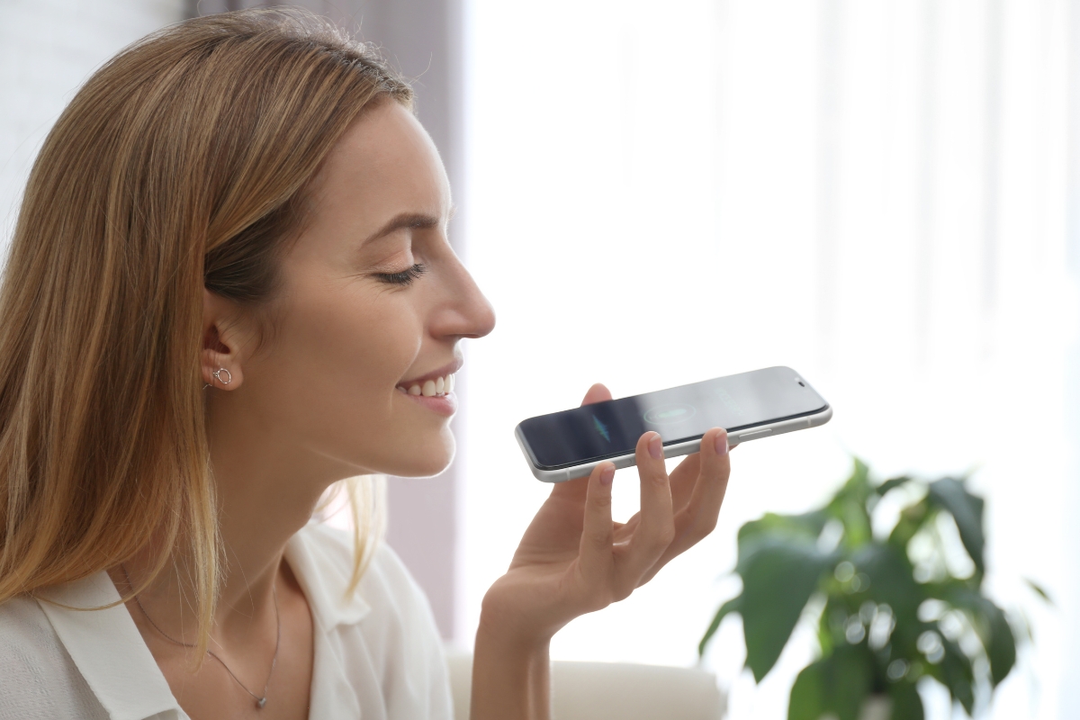 A person is holding a smartphone close to their mouth, smiling while speaking into it. Behind them, there is a blurred background with natural light and a plant, perhaps discussing tips on how to optimize your website for voice search.