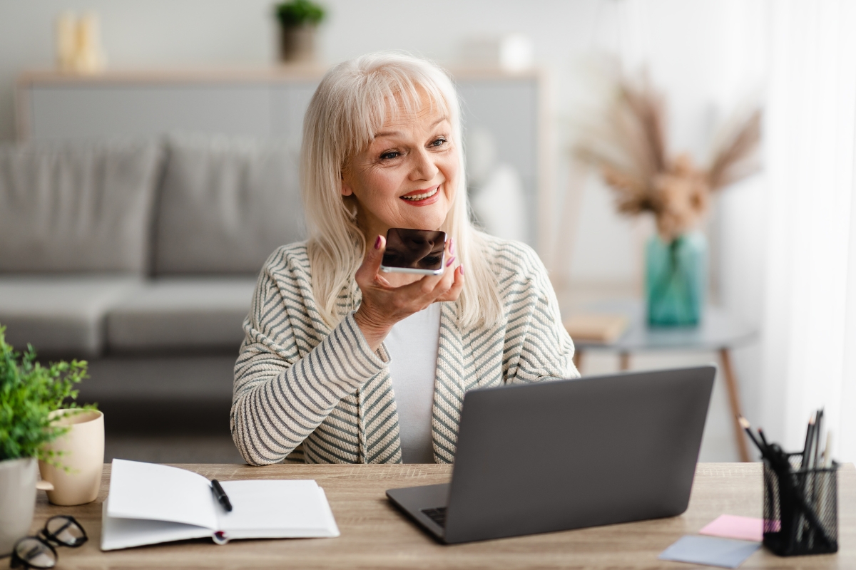 A smiling woman speaks into her phone at a desk with a laptop, notebook, and plants in a well-lit room, perhaps discussing tips on how to optimize your website for voice search.