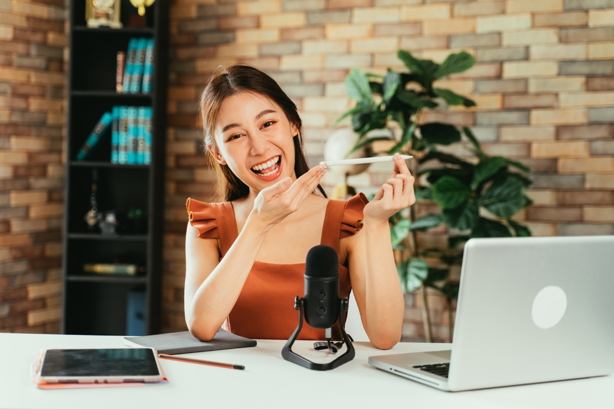 A smiling person sits at a desk with a microphone, pointing at something off-camera. In front of them are a laptop, tablet, notebook, and pencil. A bookcase and plant adorn the background as they discuss influencer marketing ROI for small businesses.