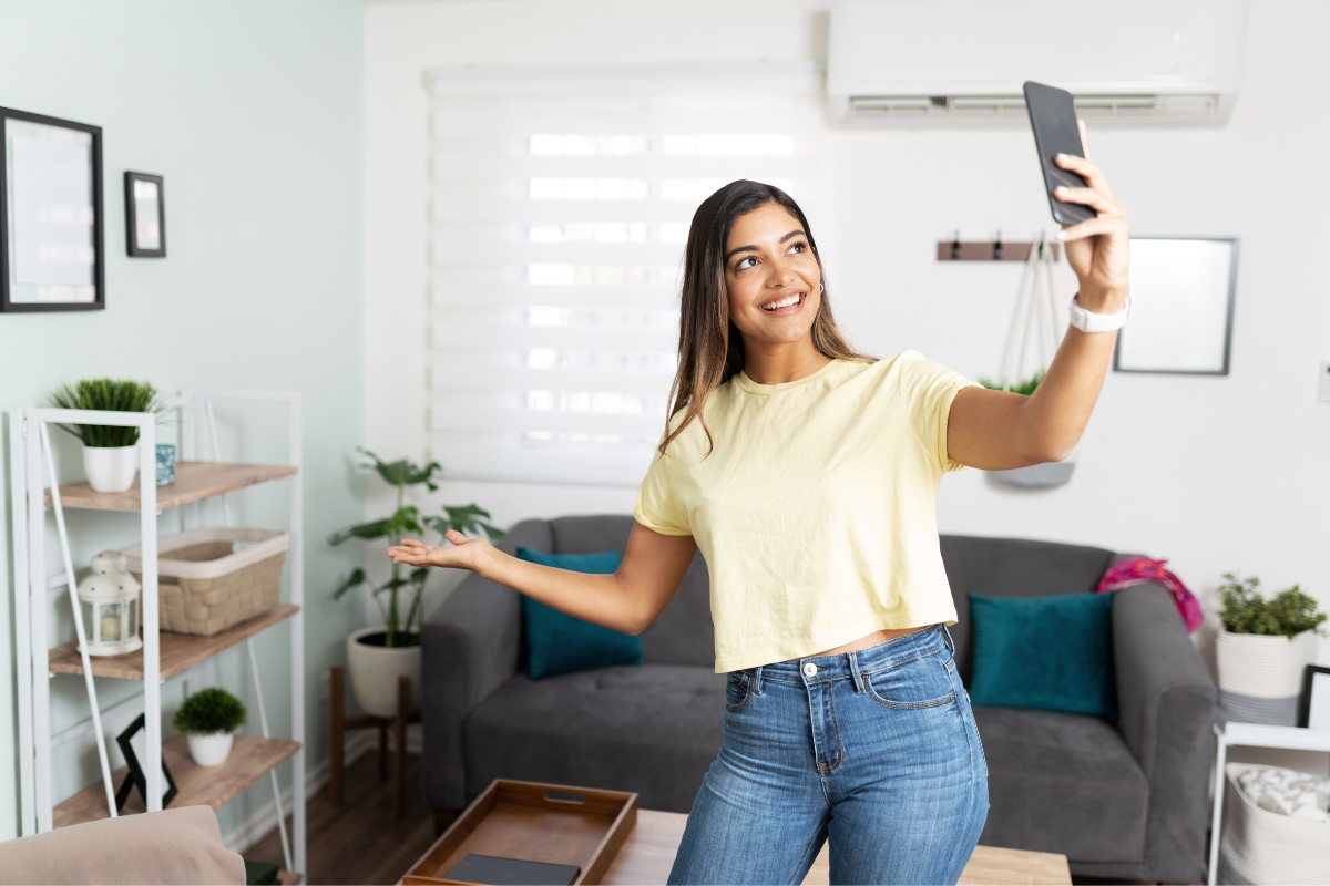 A person stands in a living room, smiling and holding a smartphone for a selfie. The room is decorated with plants, shelves, and a gray sofa. It's the perfect setting for an influencer marketing ROI snapshot.