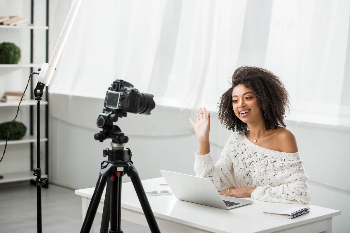 A woman sits at a desk with a laptop, waving at a camera on a tripod while recording content to boost influencer marketing ROI.