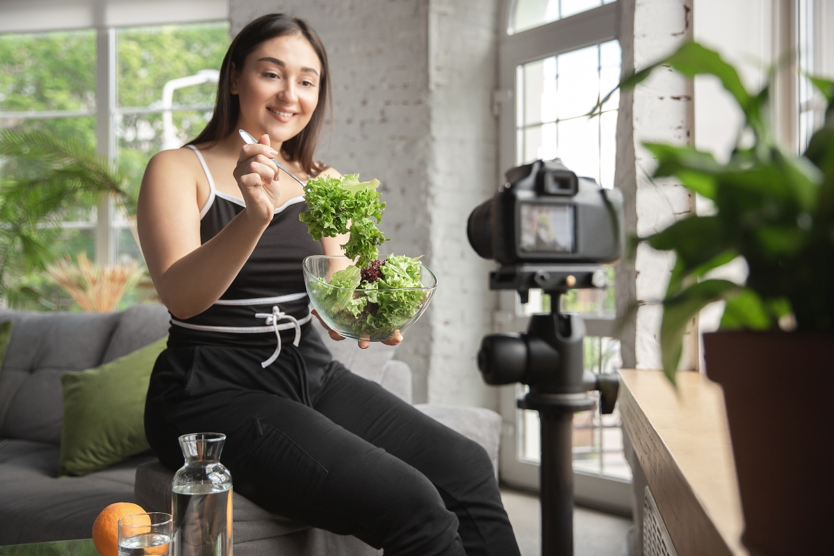 A woman sits on a couch holding a bowl of salad while recording herself with a camera on a tripod, illustrating influencer marketing ROI in real time.