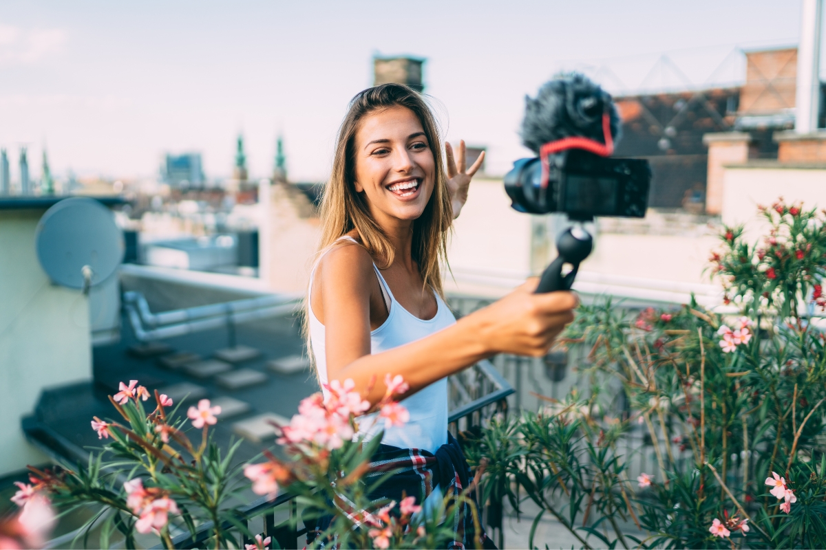 A woman smiles and waves while recording herself on a camera mounted on a handheld stabilizer, standing on a rooftop with pink flowers in the foreground, demonstrating how influencer marketing ROI can be visually appealing and effective for small businesses.