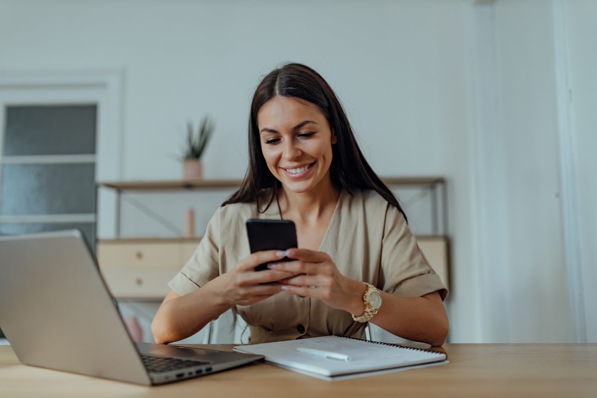 A person with long hair is sitting at a desk, smiling while using a smartphone to look up LinkedIn Marketing Tips. A laptop, notebook, and pen are also on the desk. A shelf with a plant is in the background.