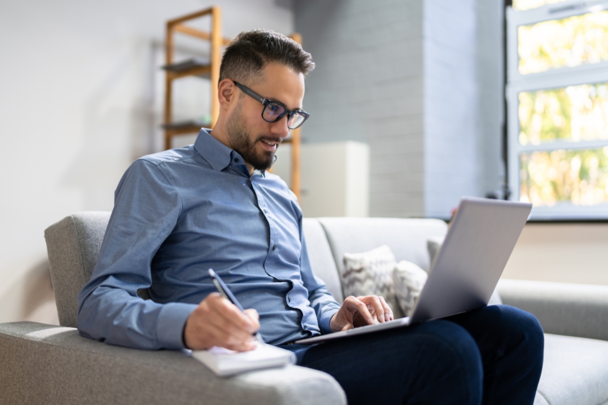 A man wearing glasses and a blue shirt sits on a couch, meticulously working on a laptop and taking notes with a pen and notepad, perhaps jotting down some LinkedIn marketing tips.