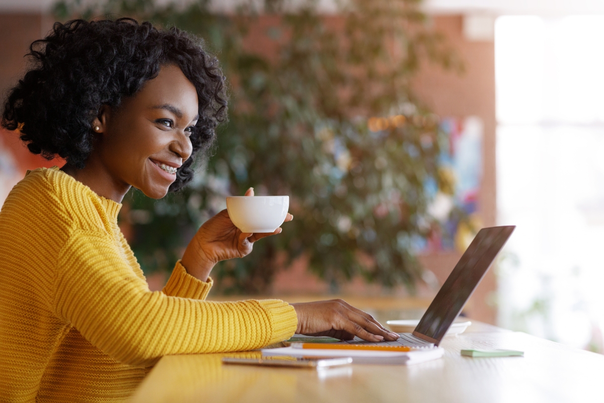 A person in a yellow sweater sits at a desk, holding a cup of coffee in one hand and typing on a laptop with the other. They're smiling and looking at the screen, likely discovering some useful LinkedIn Marketing Tips.