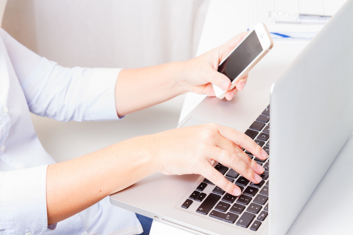 A person is using a laptop and holding a smartphone, focusing on the hands. The individual seems engaged in LinkedIn Marketing Tips in a white, bright setting with office supplies in the background.