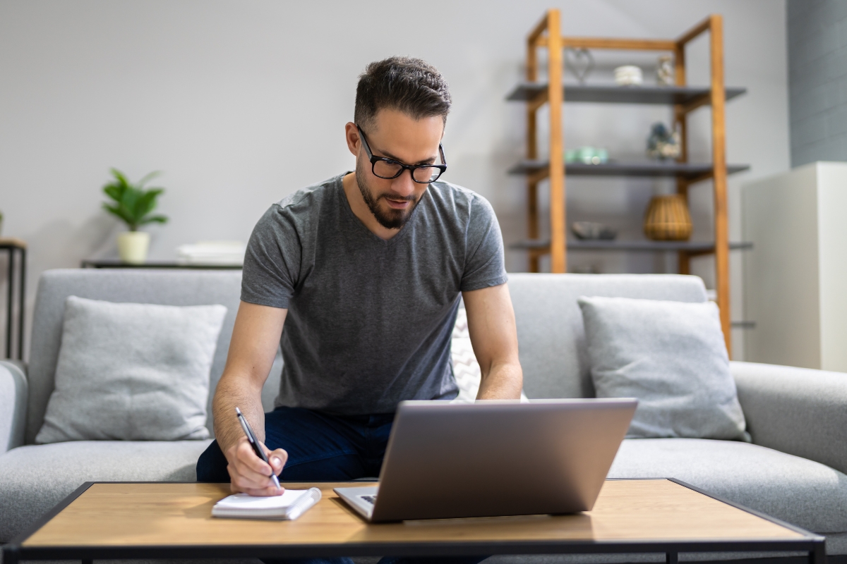 A man wearing glasses and a gray t-shirt is sitting on a sofa, taking notes on LinkedIn marketing tips in a notepad while using a laptop on the coffee table in front of him.