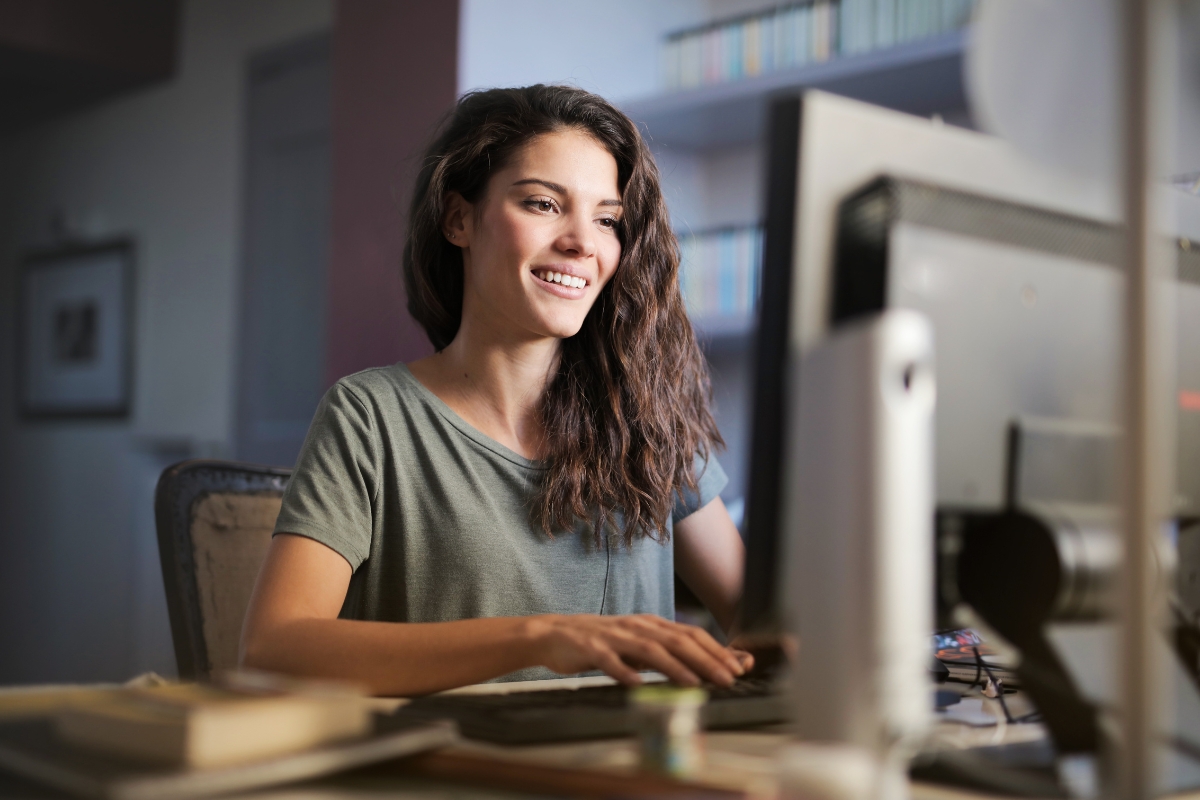 A woman with long, wavy hair smiles while typing on a computer in a room with bookshelves in the background, perhaps working on LinkedIn Marketing Tips.