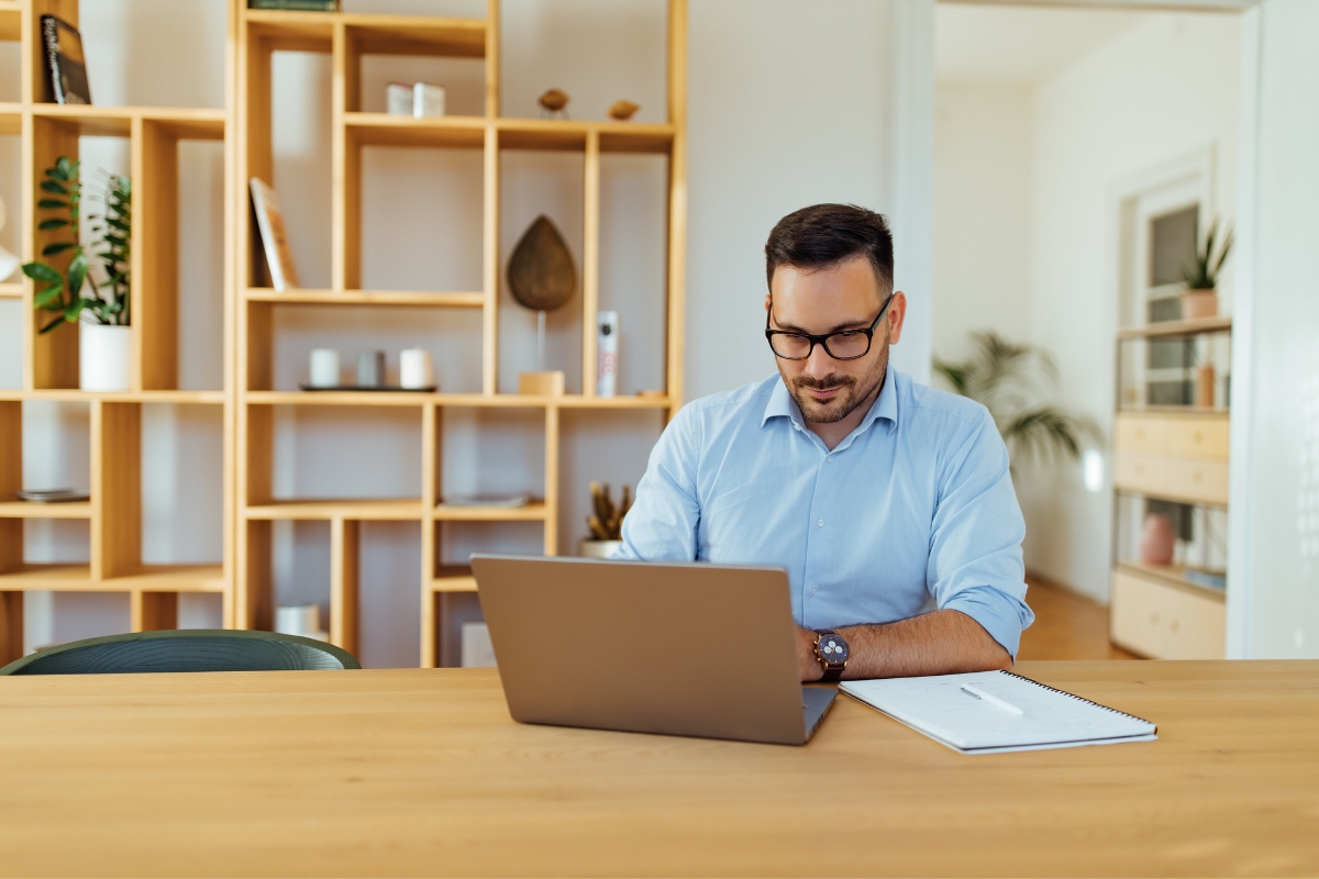 A man wearing glasses and a light blue shirt is sitting at a wooden table, working on a laptop with a notebook labeled "LinkedIn Marketing Tips" beside him. Behind him is a wooden shelving unit with various items and plants.