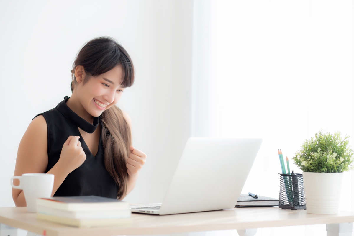 A woman in a black top, smiling and holding clenched fists, sits at a desk with a laptop, books, LinkedIn marketing tips printed on notes, a cup, and a potted plant.