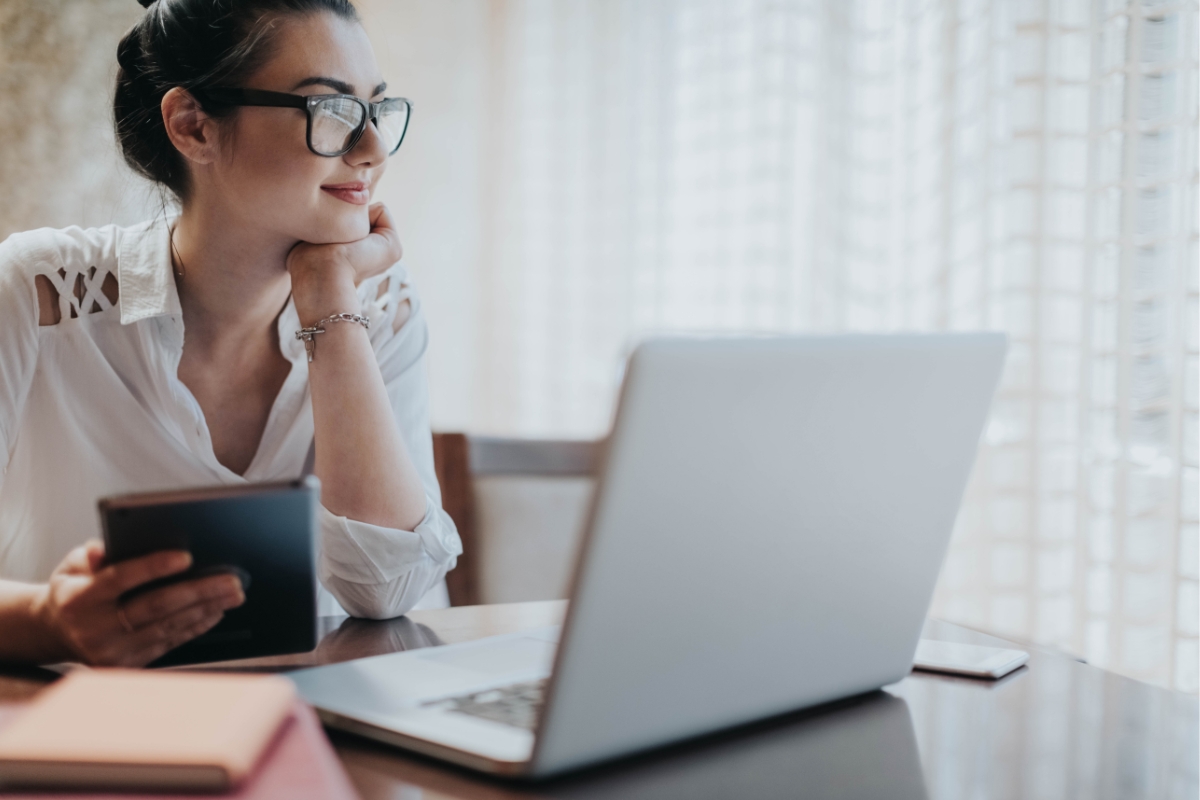 A person wearing glasses and a white shirt is sitting at a table, holding a tablet, and looking at a laptop screen, possibly exploring LinkedIn Marketing Tips.