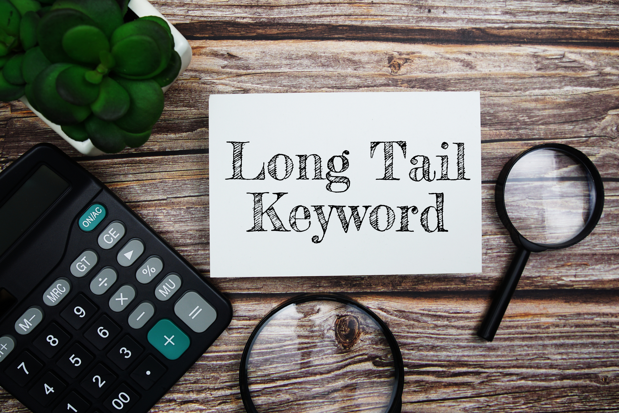 An index card with "Long Tail Keywords" text rests on a wooden table, accompanied by a calculator, magnifying glass, and a small potted plant.