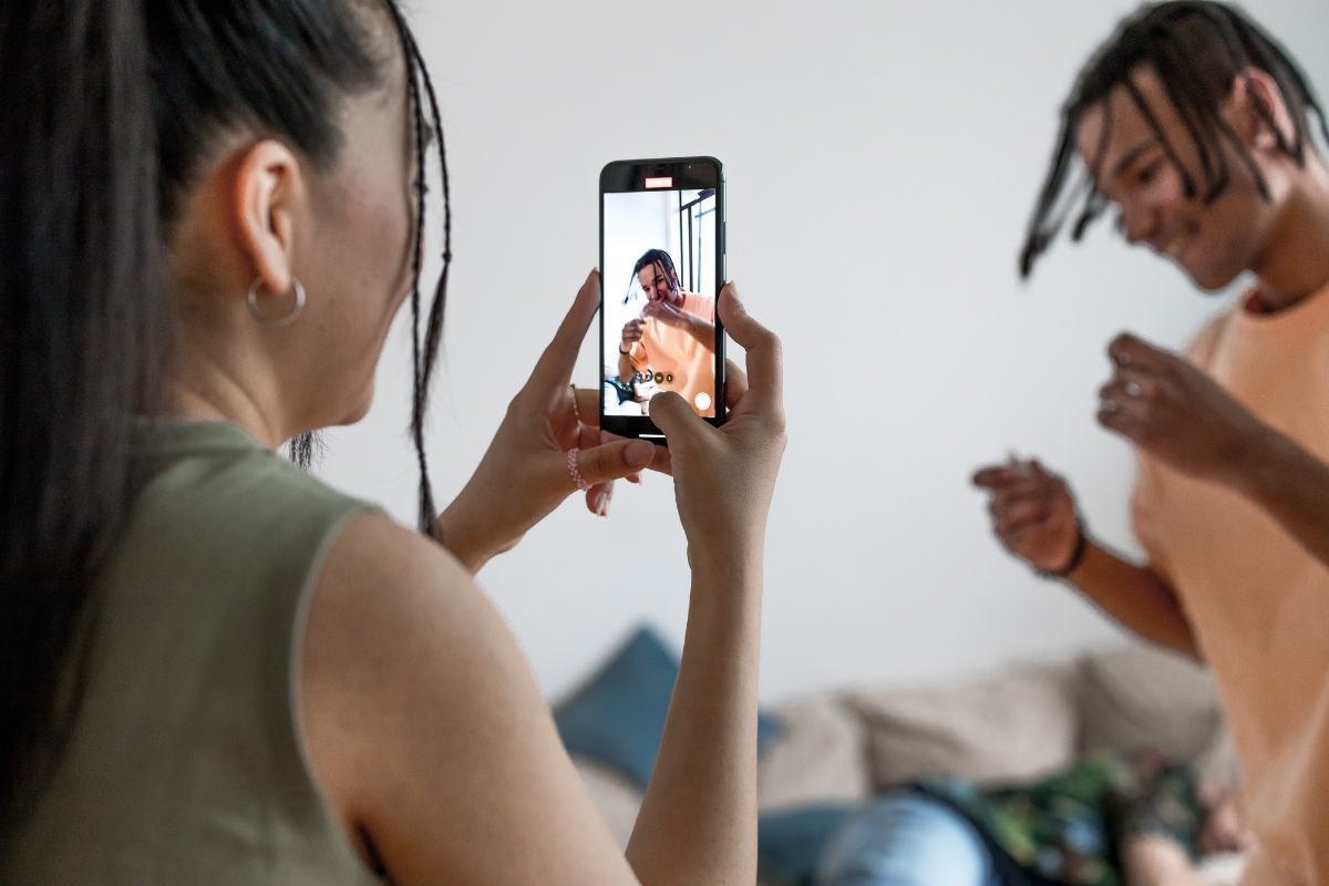 A woman takes a photo of a man using a smartphone. The man, who is smiling and has braided hair, is standing in the background and wearing a peach-colored shirt, capturing moments for marketing with Instagram Reels.