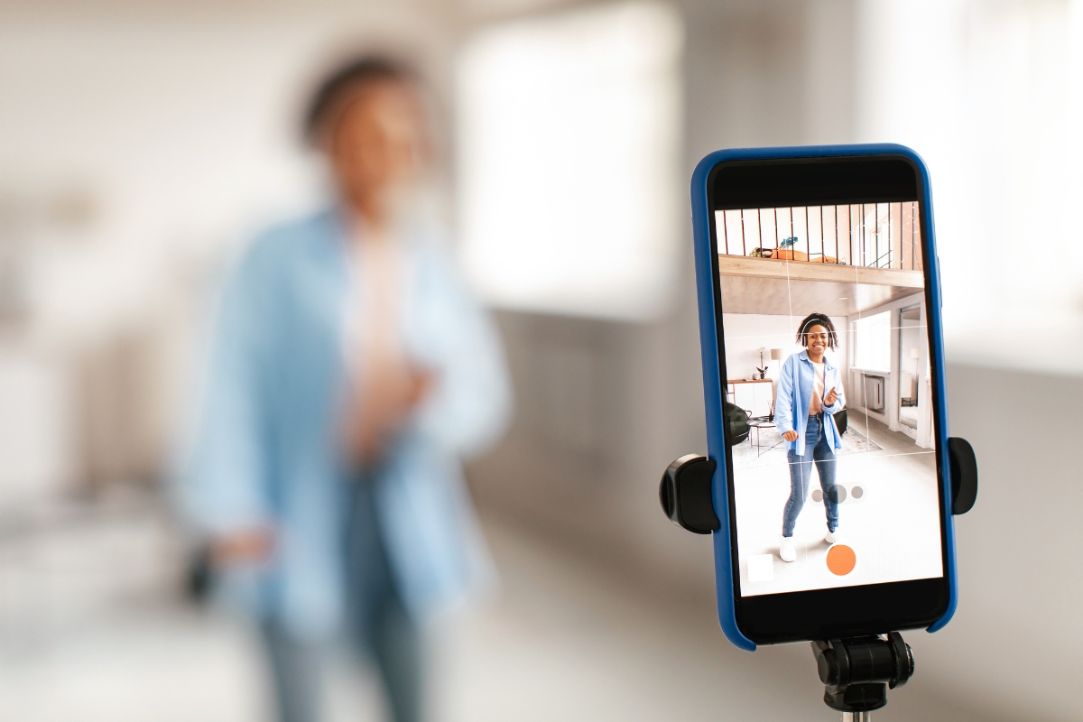 A smartphone on a tripod records a person in a blue shirt and jeans inside a bright room. The person is blurred in the background, while the phone screen shows a clearer image of them posing for an Instagram Reels marketing campaign.