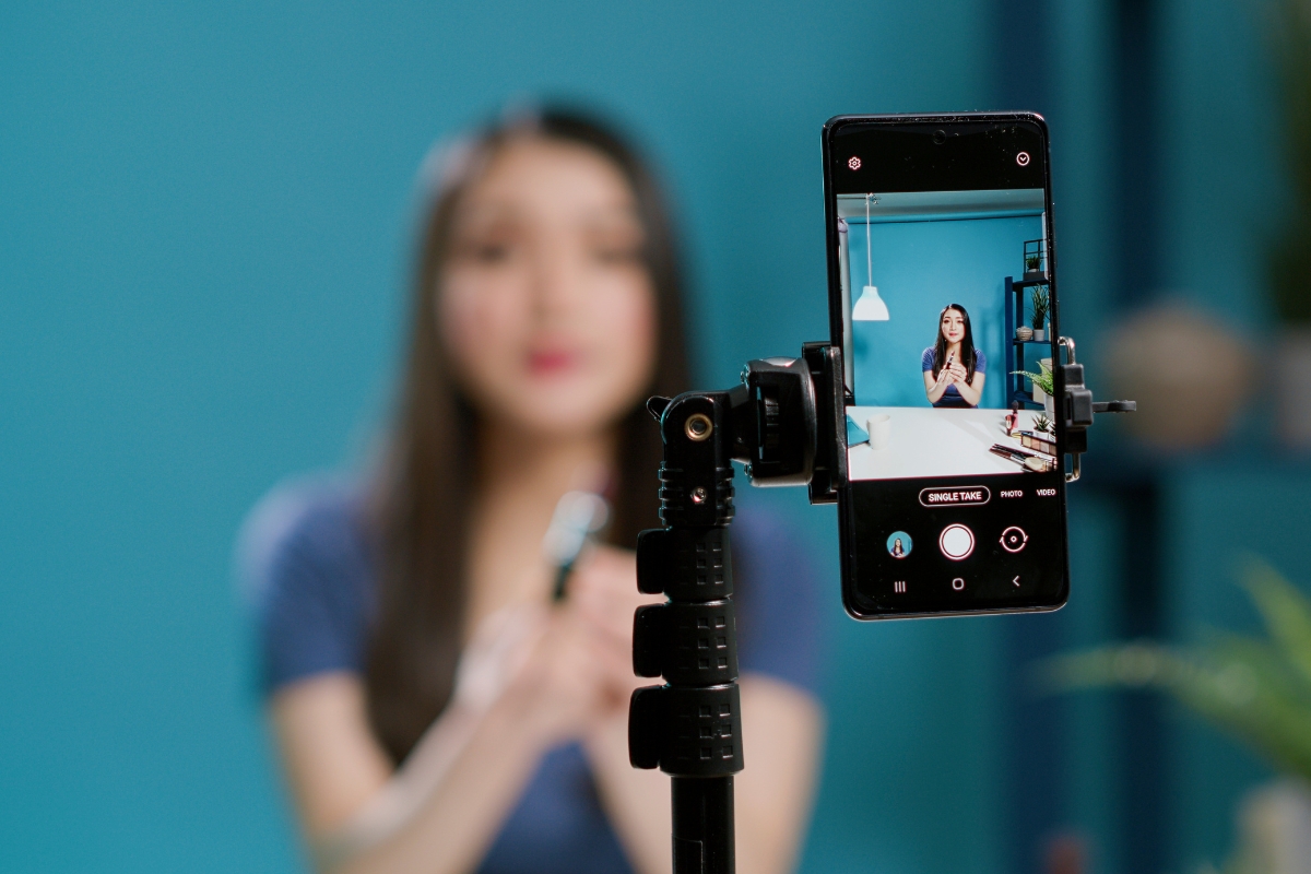 A woman in a blue shirt is being recorded by a smartphone mounted on a tripod with a blue background, creating content for Marketing with Instagram Reels.