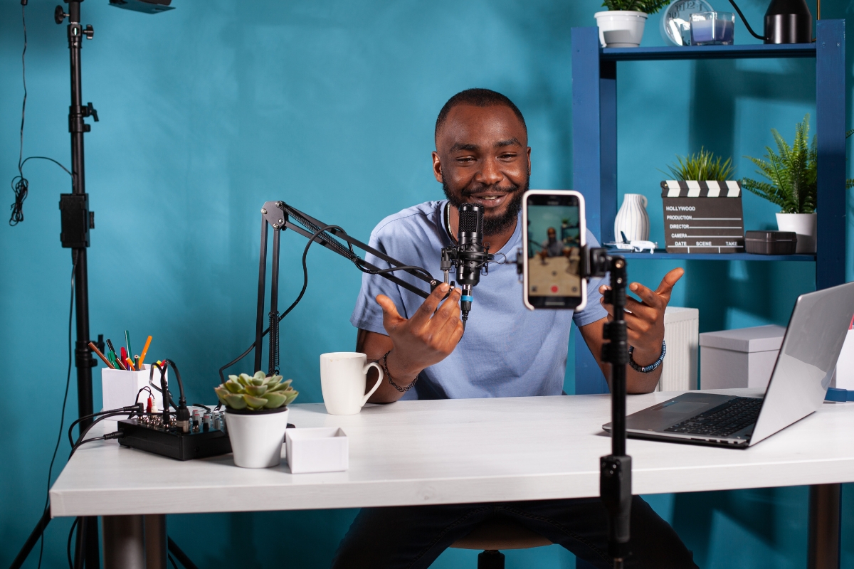 A man sits at a desk in a recording setup with a microphone, smartphone on a stand, laptop, and sound mixer. He speaks energetically about marketing with Instagram Reels, gesturing with his hands to emphasize his points.