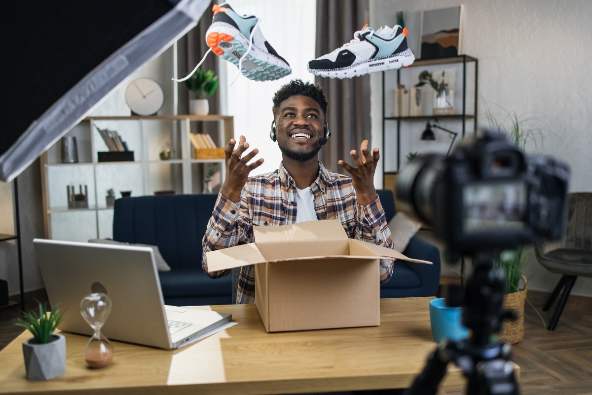 A person sitting at a desk with a laptop, unboxing a pair of sneakers in front of a camera, smiling, with one sneaker in each hand and an opened shipping box in front—perfect for marketing with Instagram Reels.