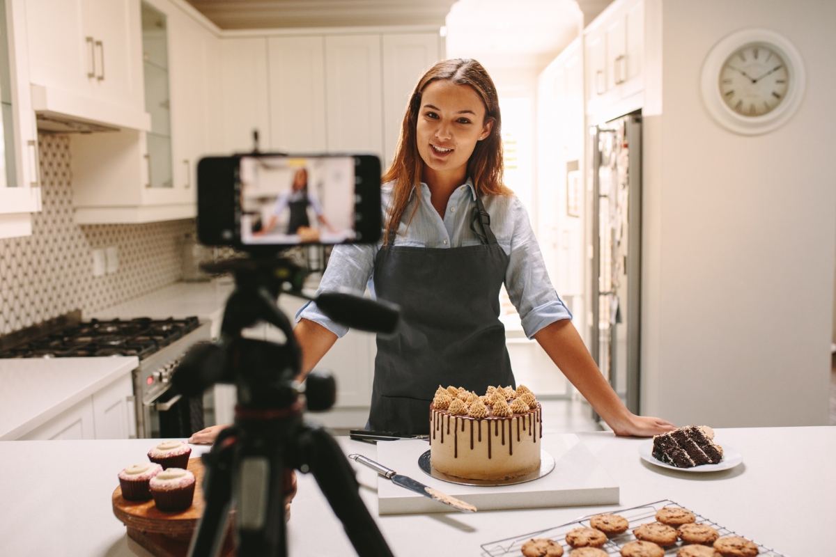 A woman in a kitchen records herself with a smartphone, skillfully using Instagram Reels for marketing. She stands behind a decorated cake, alongside cupcakes and cookies laid out on the countertop.