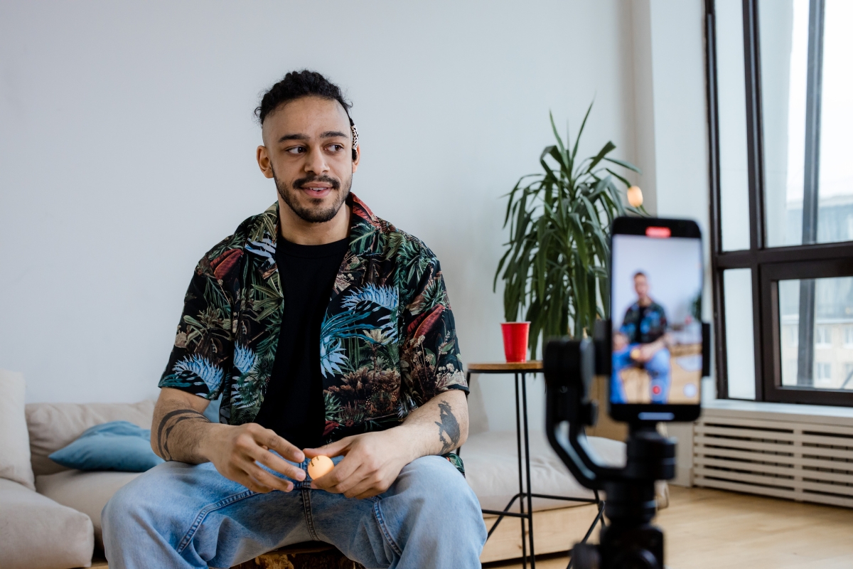 A man sits in a decorated shirt and jeans, holding an object while recording himself with a smartphone on a tripod, likely creating content for marketing with Instagram Reels. A plant and red cup are in the background.