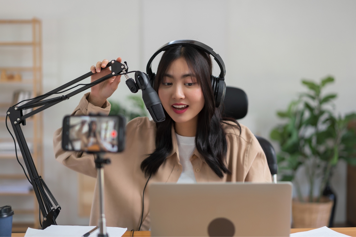 A person wearing headphones speaks into a microphone while sitting at a desk with a laptop, marketing with Instagram Reels, and filming themselves on a smartphone.