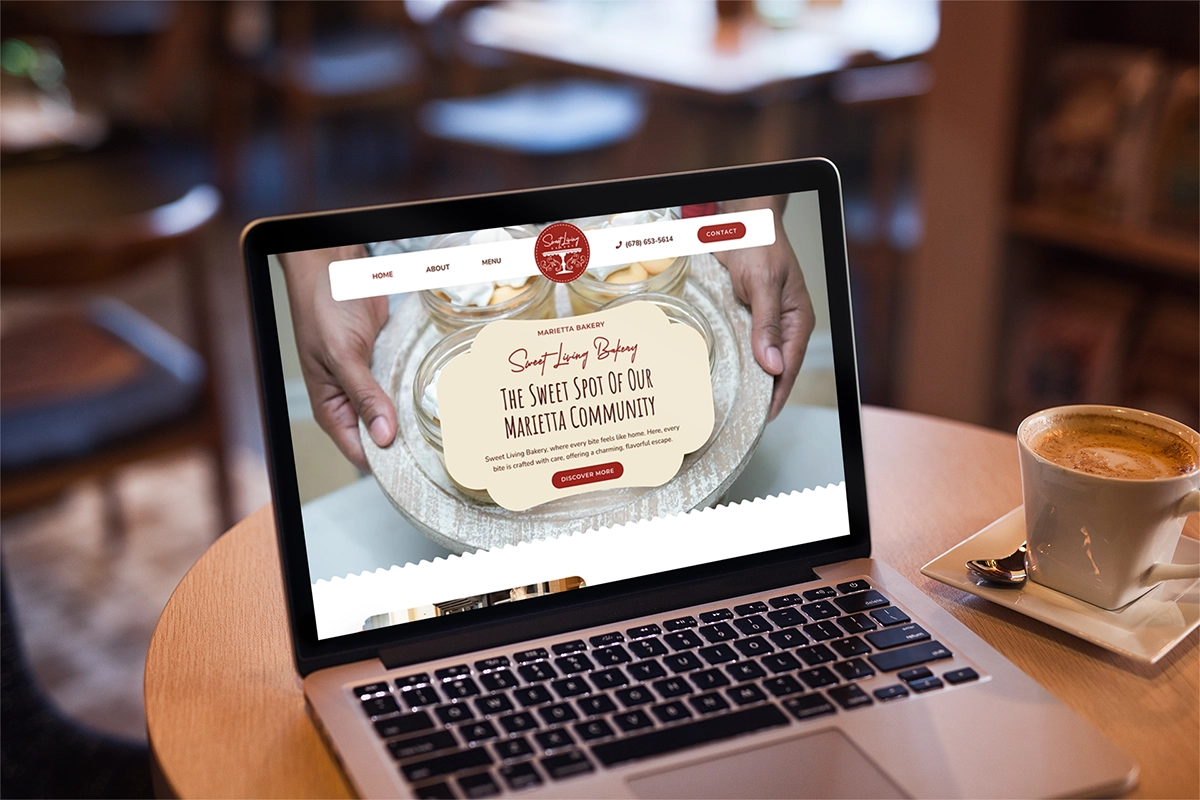 A laptop displaying a bakery website with the text "Sweet Apron Bakery: The Sweet Spot of Our Marietta Community" rests on a table next to a cup of coffee and saucer, capturing the essence of sweet living among local delights.