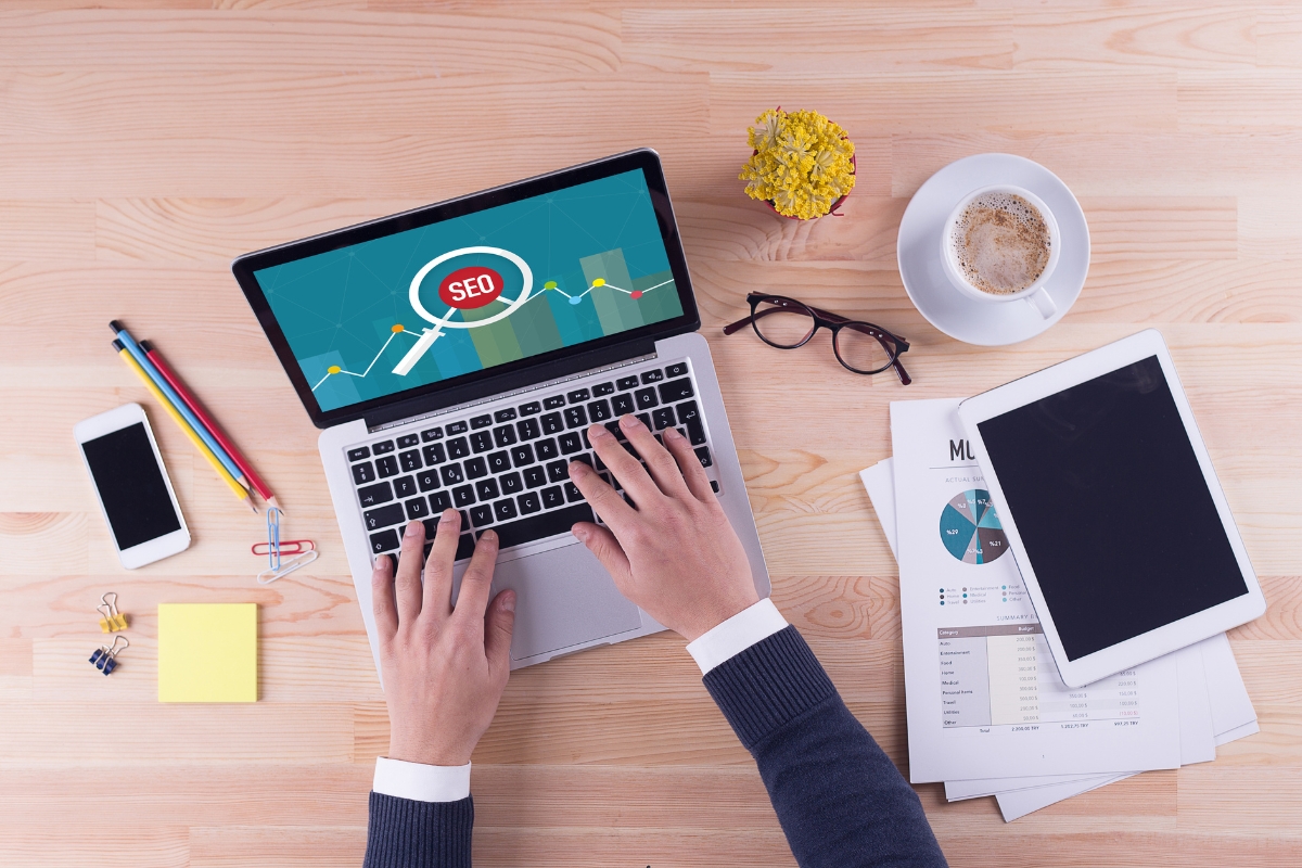 A person works on a laptop displaying a programmatic SEO screen. The desk has a phone, tablet, charts, glasses, a cup of coffee, pens, and sticky notes.