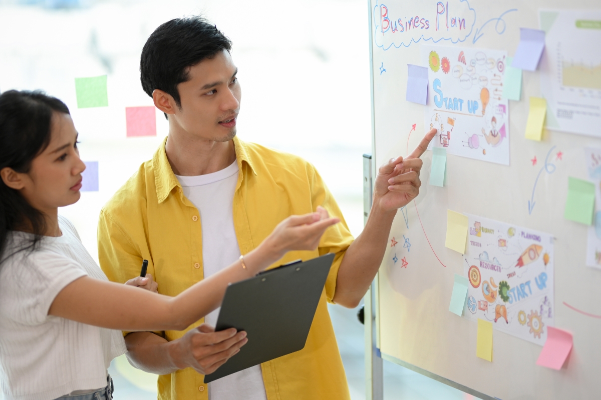 Two individuals, one holding a clipboard, discuss a real-time marketing plan in front of a whiteboard covered with colorful diagrams and notes.