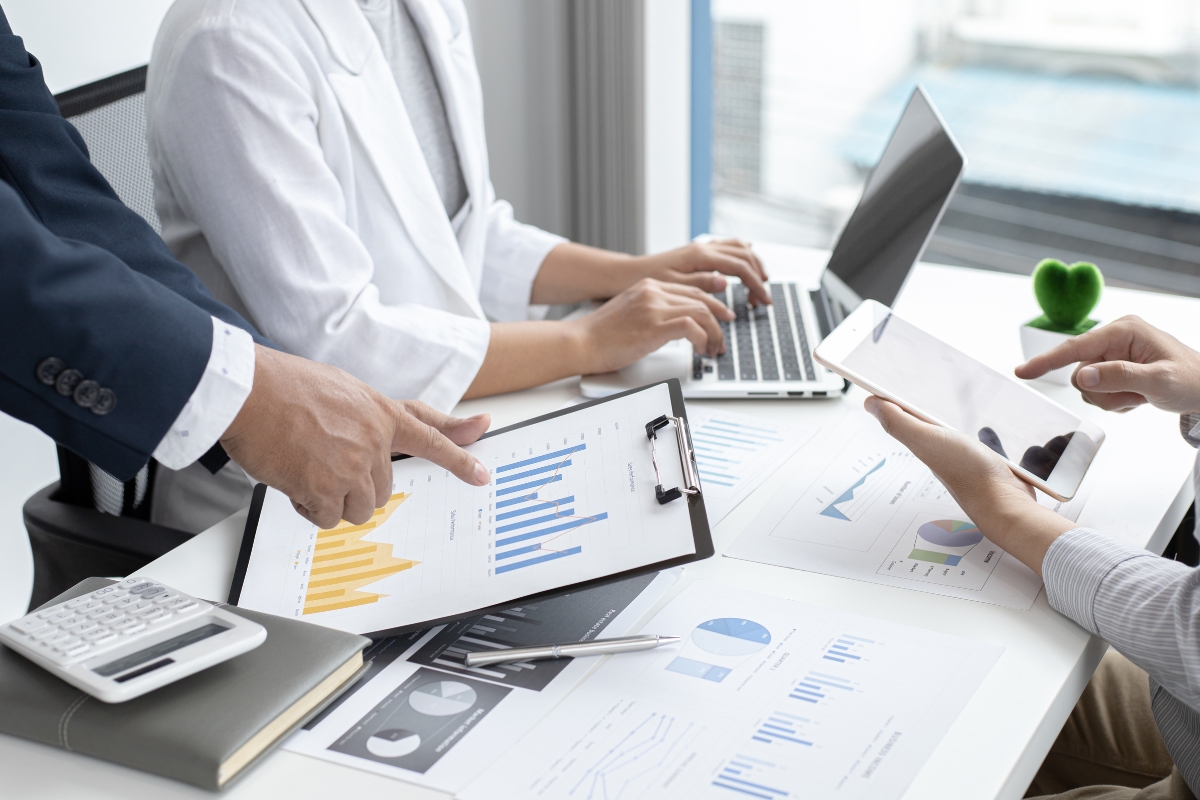 Three people in an office work with charts and graphs on paper, a tablet, and a laptop. One person points to a graph on a clipboard while the others review real-time marketing data on their devices.