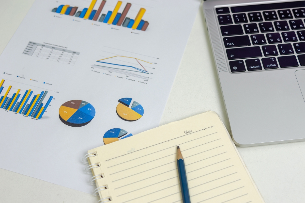 A paper with bar charts, pie charts, and a line graph next to an open notebook with a pencil and a laptop keyboard, illustrating real-time marketing dynamics.