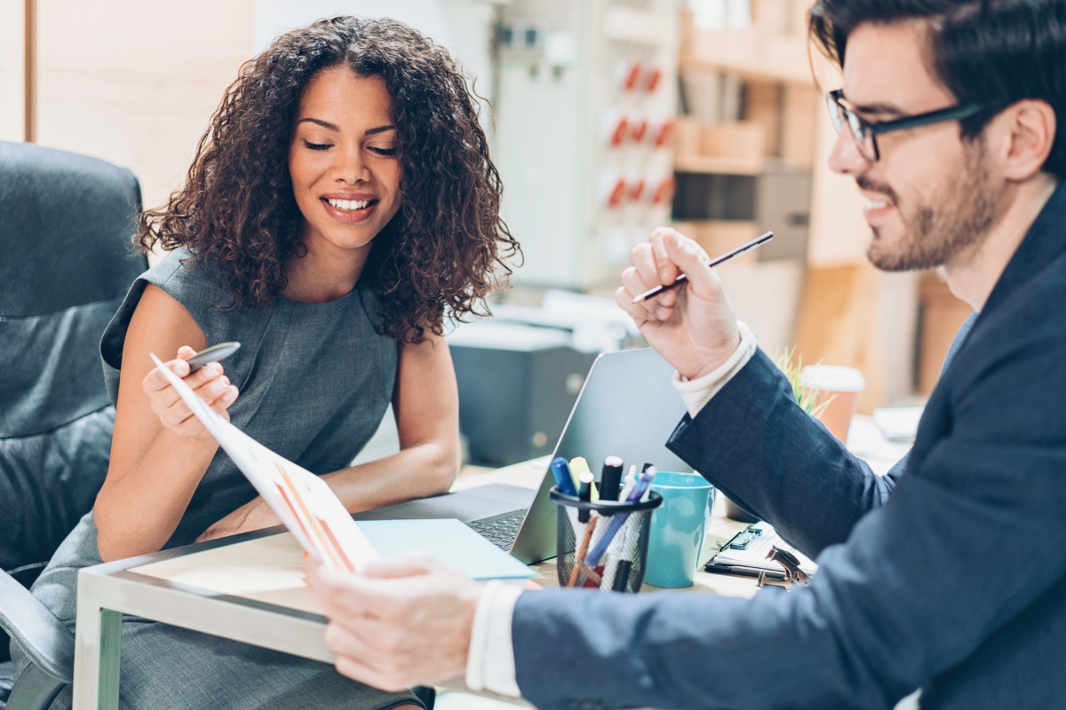 Two colleagues sit at a desk in an office. The woman is smiling and holding a document related to real-time marketing. The man is holding a pen and looking at the document. A laptop and office supplies are on the desk.