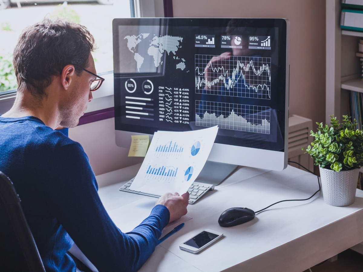 A person in a blue shirt sits at a desk, analyzing charts and graphs on paper and a computer screen displaying various data visualizations from SEO audit tools. A plant, phone, and mouse are on the desk.