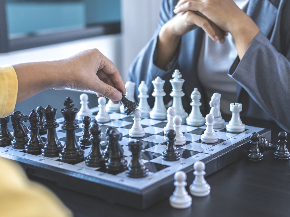 Two people are engaged in a chess game, with one person moving a black knight piece. Both are seated at a table, intensely focused on the chessboard, much like strategists diving into SEO competitive analysis.