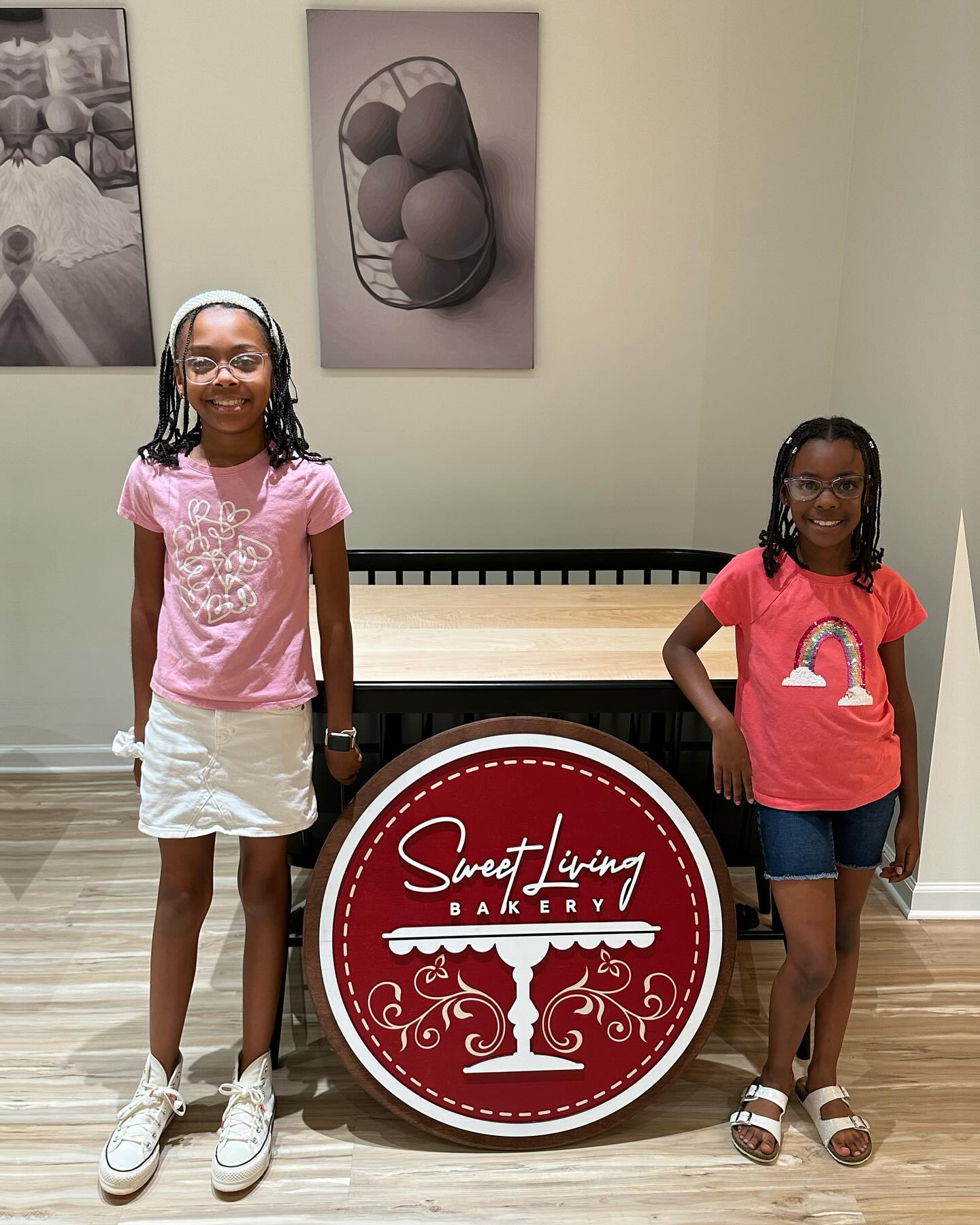 Two girls smiling and standing beside a round sign that reads "Sweet Living Bakery" in a well-lit room with framed wall art.