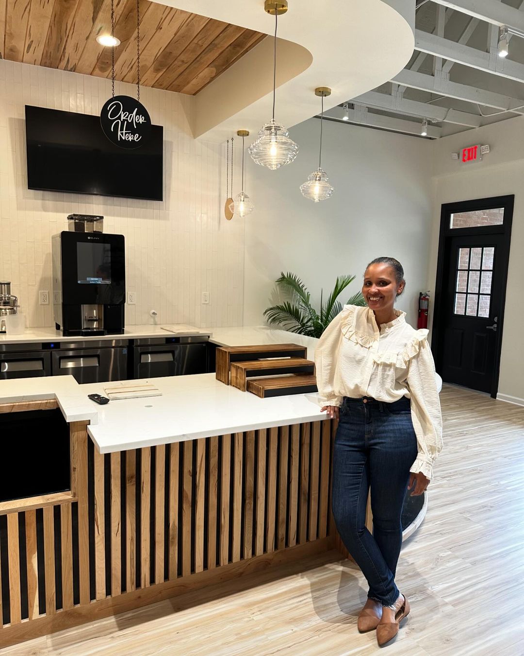 The woman stands smiling next to a counter in the modern, well-lit Sweet Living Bakery with a wooden ceiling and hanging lights. A black door and a potted plant are visible in the background.