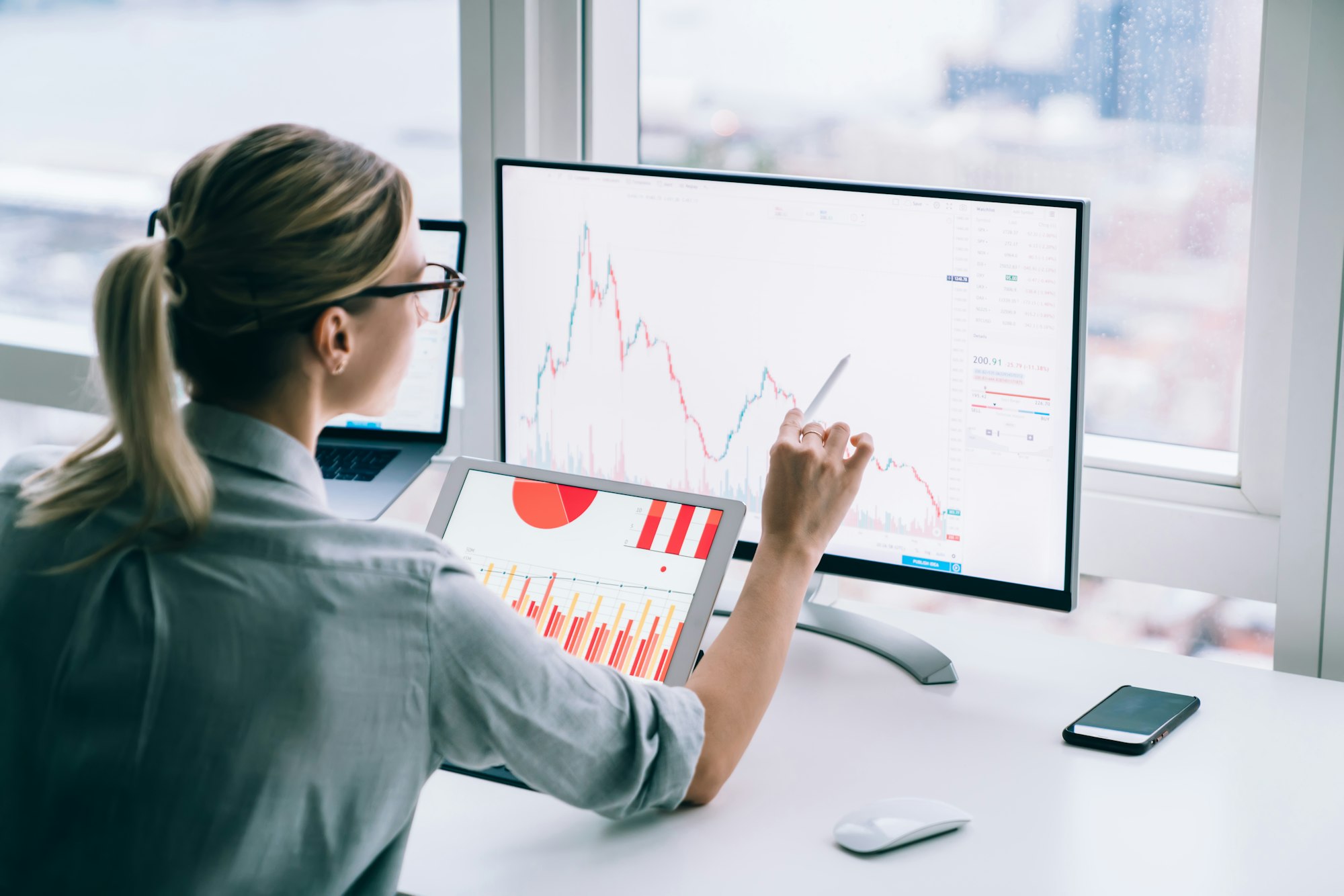 A person analyzing financial graphs on two monitors at a desk, one showing a bar chart and the other displaying a line graph.