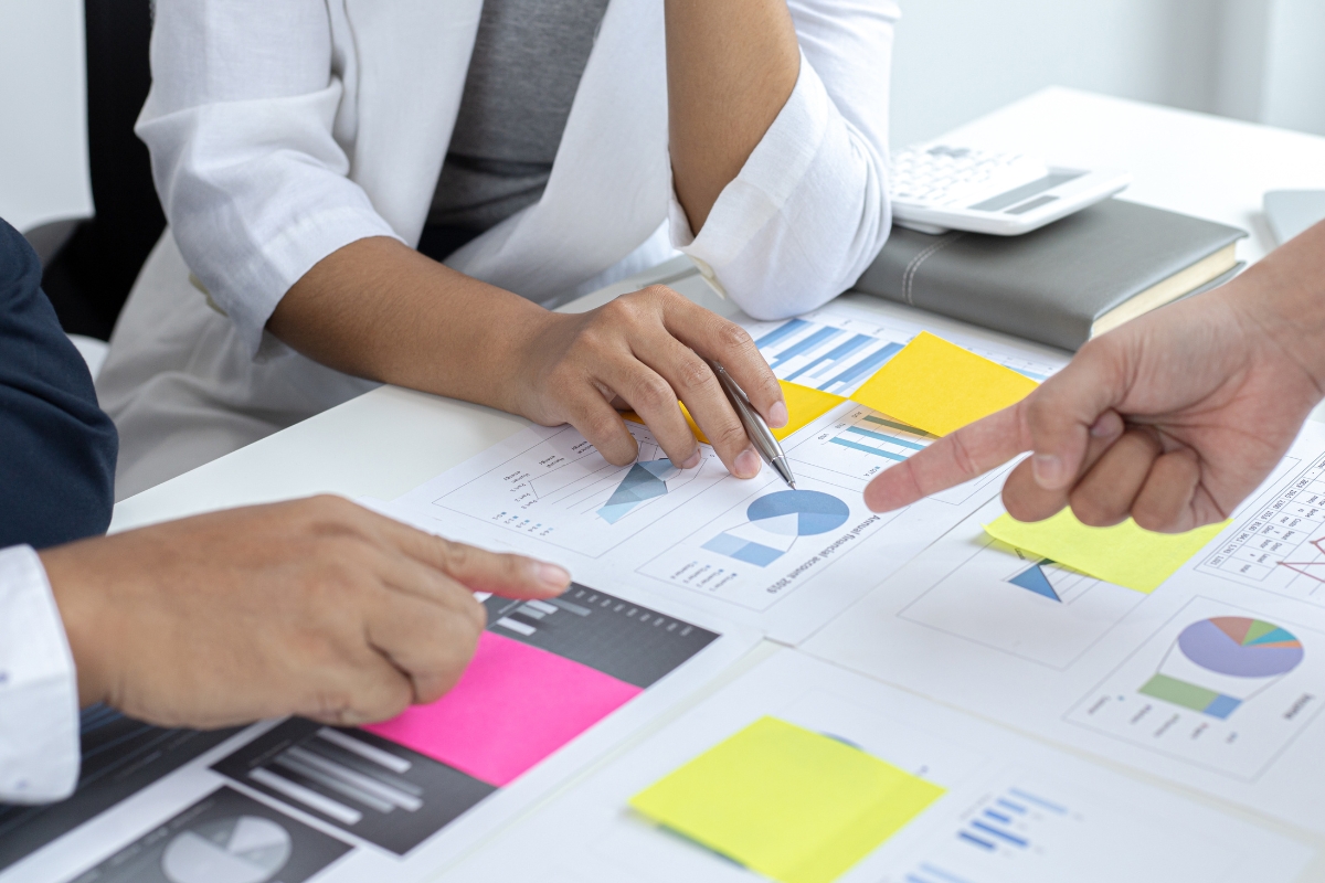 People discussing and pointing at financial charts and graphs spread out on a table, with colorful sticky notes displaying notes like "what does UTM stand for" and a calculator visible.