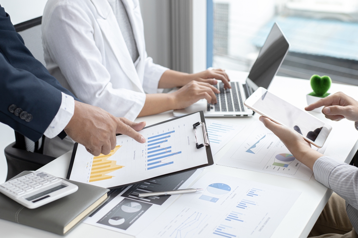 Three people working at a desk with charts, graphs, and documents. One is pointing at a chart on a clipboard while explaining what "UTM" stands for, another is using a laptop, and the third is holding a tablet.