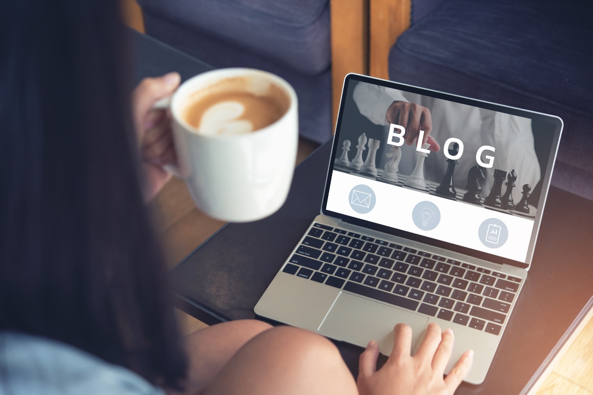 Person holding a coffee cup and using a laptop displaying a blog page that explains what is Guest Blogging, with chess pieces and icons for email, ideas, and settings shown on the screen.