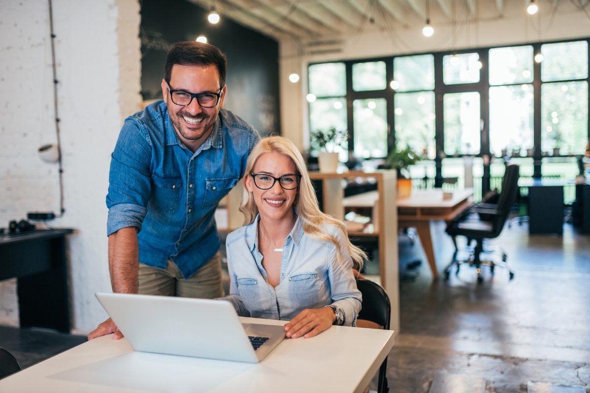 A man and a woman, both wearing glasses and denim shirts, smile in a bright office with large windows. The woman is sitting at a desk with a laptop, researching what is Guest Blogging, while the man stands beside her.