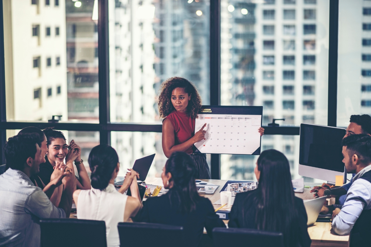 A woman stands holding a calendar, presenting "What is Guest Blogging?" to a group of colleagues seated around a conference table in an office with large windows overlooking city buildings.