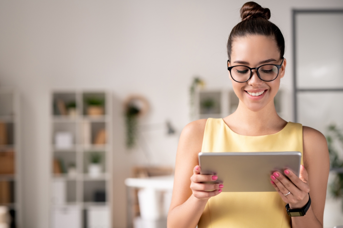 A woman in a yellow top and glasses is smiling while using a tablet in a modern office space with shelves and plants. She seems engaged, perhaps researching what is guest blogging.