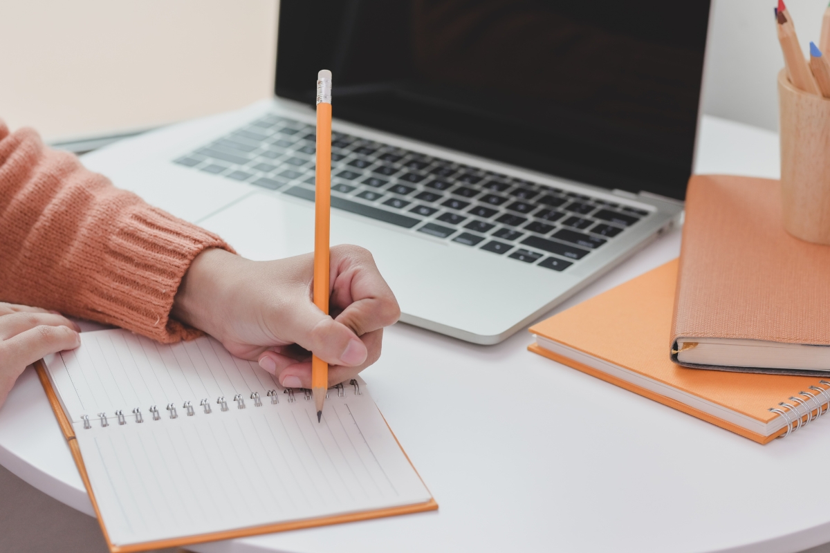 A person wearing an orange sweater is writing in a spiral notebook with a pencil on a desk that also has a laptop displaying "What is Guest Blogging?", another notebook, and a cup holding pencils.