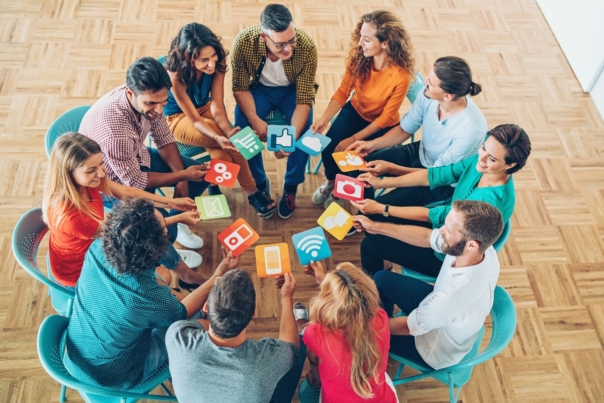 A group of people sitting in a circle on wooden flooring, holding icons of various apps and social media platforms, engaging in a discussion about why social media is important for modern communication and connectivity.