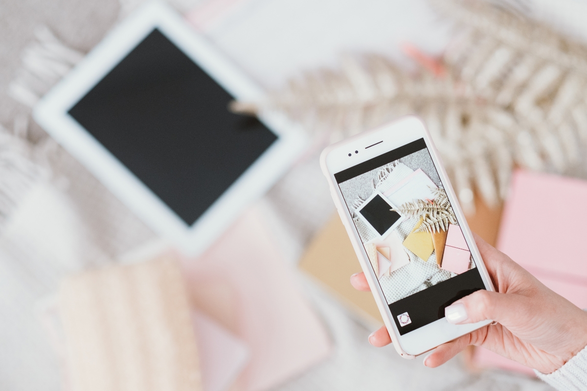 A person holds a smartphone, capturing a flat lay arrangement that includes a tablet, notebook, dried leaves, and other stationery items on a light surface. This scene subtly answers the question of why social media is important in sharing aesthetically pleasing moments and inspiration.