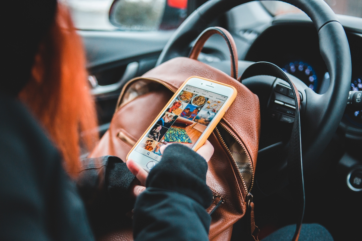 Person using a smartphone while sitting in a car, with a brown leather bag on their lap. The phone screen displays a collage of photos, illustrating why social media is important for staying connected.