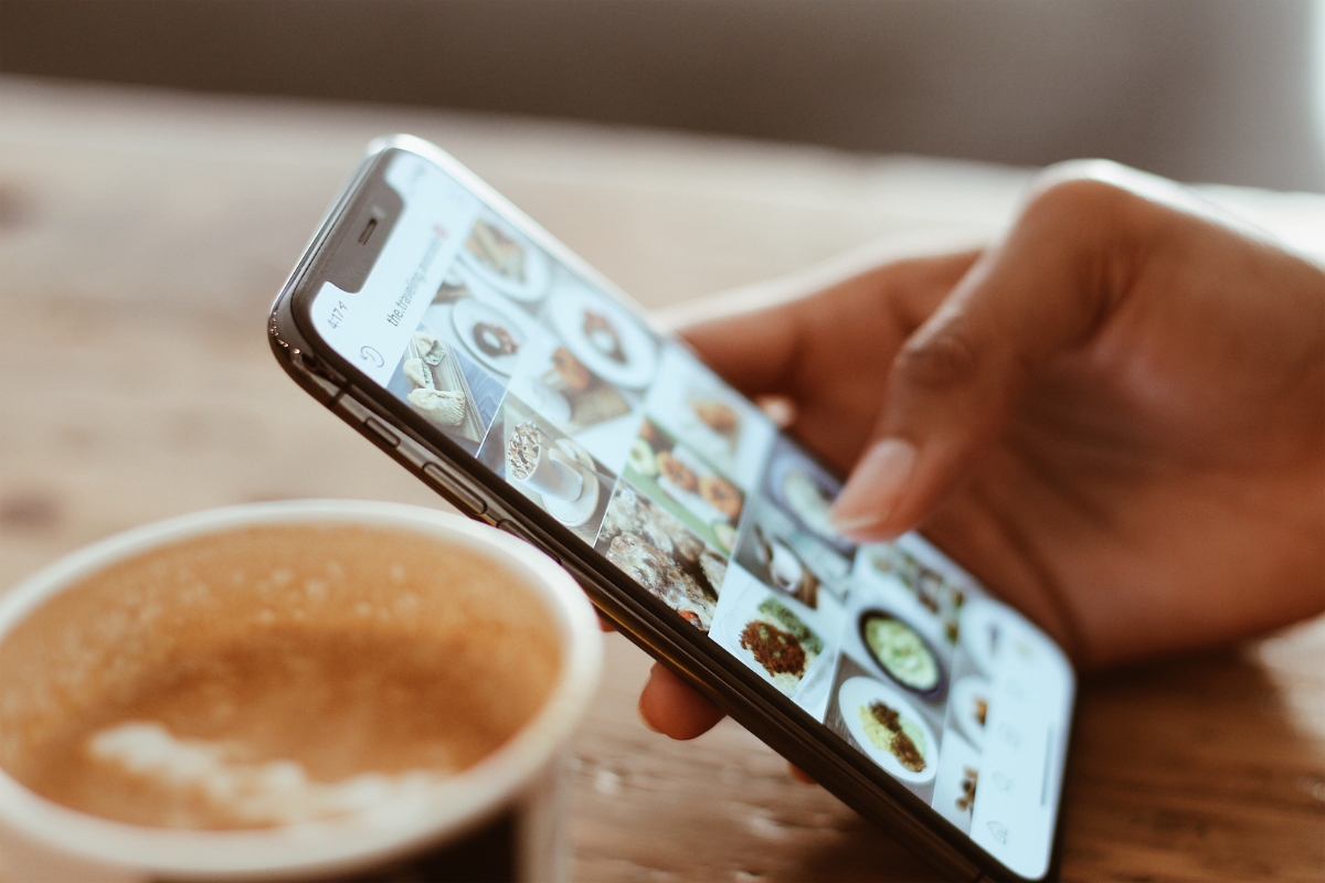 A person holds a smartphone displaying various food images next to a cup of coffee, subtly exemplifying why social media is important in sharing daily moments and culinary experiences.