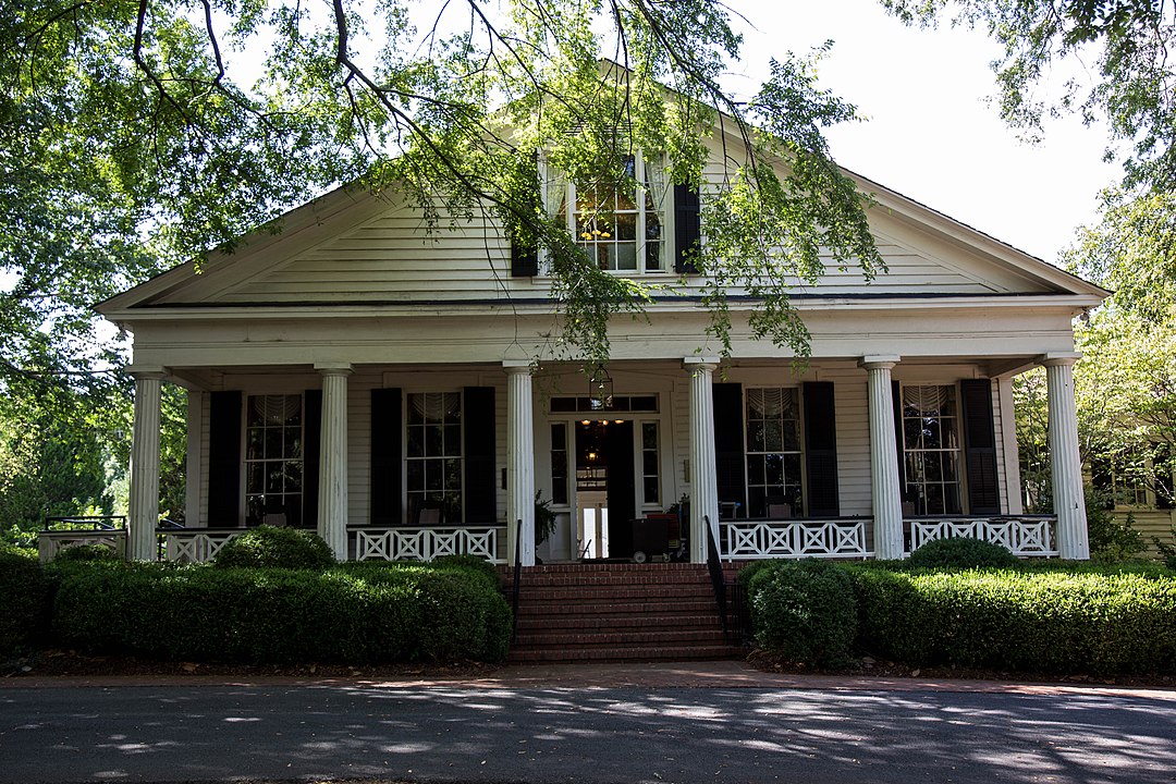 A white, one-story house with a front porch supported by six columns. The entrance is flanked by black shutters, surrounded by trees and bushes. Nestled in Marietta, this charming residence evokes the elegance of nearby Brumby Hall and its beautiful gardens.